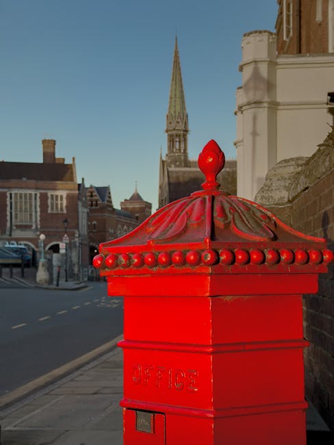 A close-up view of a bright red post box situated on a sidewalk in an urban area during daylight, with a historic church steeple and traditional brick buildings visible in the background under a clear