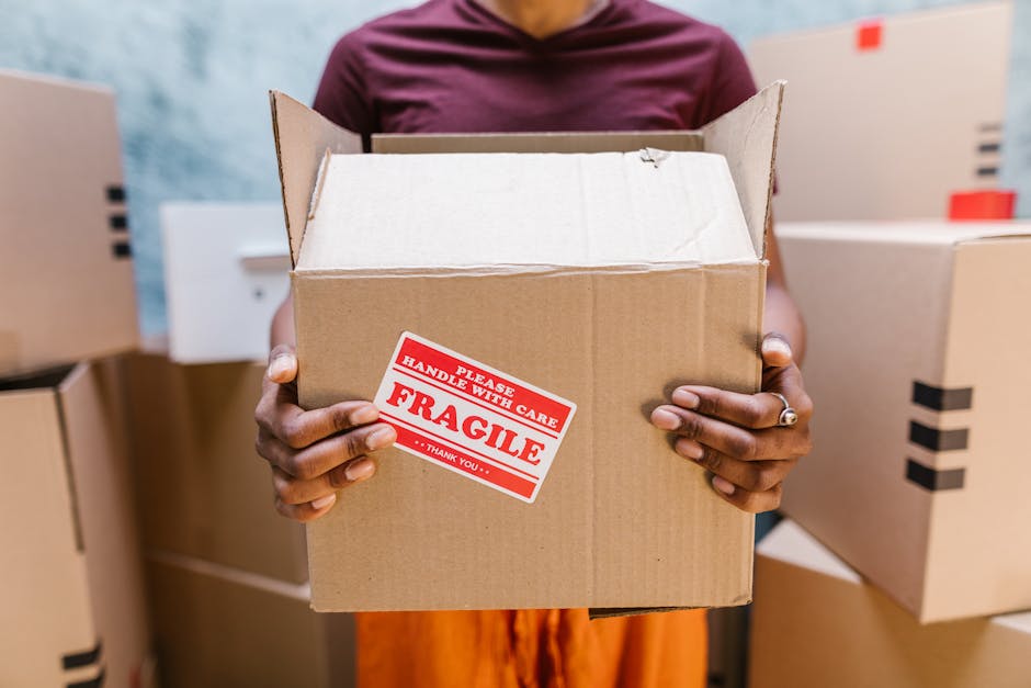 A person with dark skin is holding an open cardboard box with both hands, positioned in front of their torso in an indoor setting. The box features a red and white label that reads 'Please Handle with Care. Fragile. Thank You.' Several other closed cardboard boxes are visible in the background, some stacked and others placed on the floor or shelves. The setting appears to be a storage or packing area, with bright lighting illuminating the scene. The person is dressed in a patterned orange and black garment, and the focus is on the box and the fragile sticker, highlighting the process of packing or handling items for home relocation or moving services, as offered by Man and a Van Harrow.