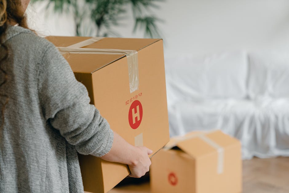 A person wearing a grey knitted sweater is using a box cutter to open a large cardboard moving box secured with clear packing tape, which features a red 'H' symbol and printed weight and size details. The scene is set indoors on a wooden floor, with a white couch draped in a fabric cover visible in the background. Multiple other closed cardboard boxes of various sizes, also sealed with tape, are arranged nearby, indicating a home relocation process. The person's hands are focused on carefully cutting the tape to access the contents inside the box. Man and a Van Harrow, a professional removals company, is likely involved in the furniture transport and packing and moving services, assisting with the efficient loading process of household items. The lighting appears natural, highlighting the details of the packaging and the ongoing packing activities related to house removals.