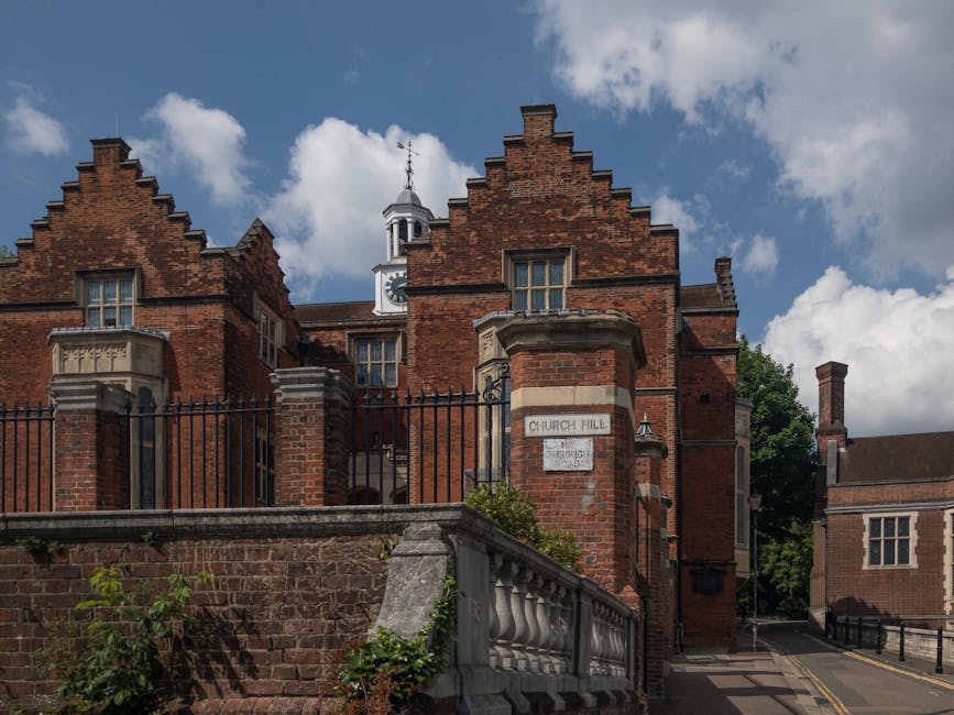 An exterior view of the historic church hall building in Harrow, featuring red brick walls with decorative stepped gables and tall sash windows. The building is surrounded by a black wrought iron fence and a stone balustrade at the street level, with a small sign reading 'Church Hall' attached to the brick wall. A narrow sidewalk runs along the front, with the sky above partly cloudy, indicating daytime. In the background, a clock tower can be seen at the top of the building, and a side street extends behind the main structure. The scene is calm and typical of a residential or community area in Harrow, suitable for house removals or furniture transport services like those provided by Man and a Van Harrow.