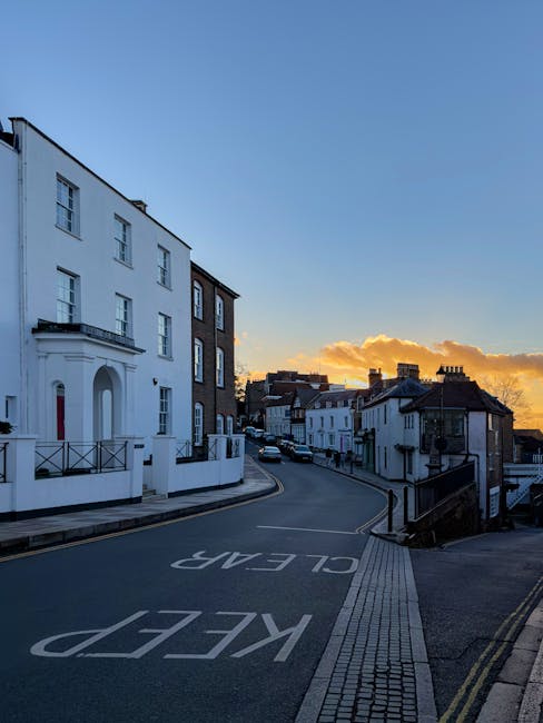 A quiet street scene in Harrow on the Hill during early evening, featuring a row of residential buildings with white and brick facades, some with small balconies and decorative moldings. The street curves gently to the right, with a paved pedestrian walkway and tactile paving along the roadside. A clear blue sky transitions to warm orange and yellow hues near the horizon, with a few scattered clouds illuminated by the setting sun. Vehicles are parked along the curb, and the road is marked with the words 'KEEP CLEAR' painted in white on the asphalt. This scene illustrates the typical urban environment where home relocation and furniture transport services might operate, and it aligns naturally with the context of house removals near Harrow School, as offered by Man and a Van Harrow.