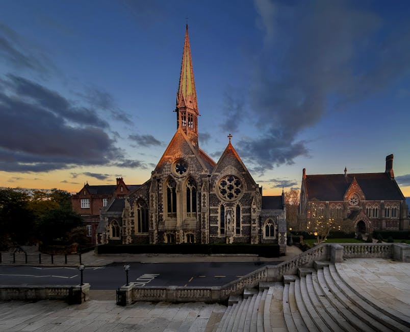 An exterior view of the historic church hall building in Harrow, featuring red brick walls with decorative stepped gables and tall sash windows. The building is surrounded by a black wrought iron fence and a stone balustrade at the street level, with a small sign reading 'Church Hall' attached to the brick wall. A narrow sidewalk runs along the front, with the sky above partly cloudy, indicating daytime. In the background, a clock tower can be seen at the top of the building, and a side street extends behind the main structure. The scene is calm and typical of a residential or community area in Harrow, suitable for house removals or furniture transport services like those provided by Man and a Van Harrow.