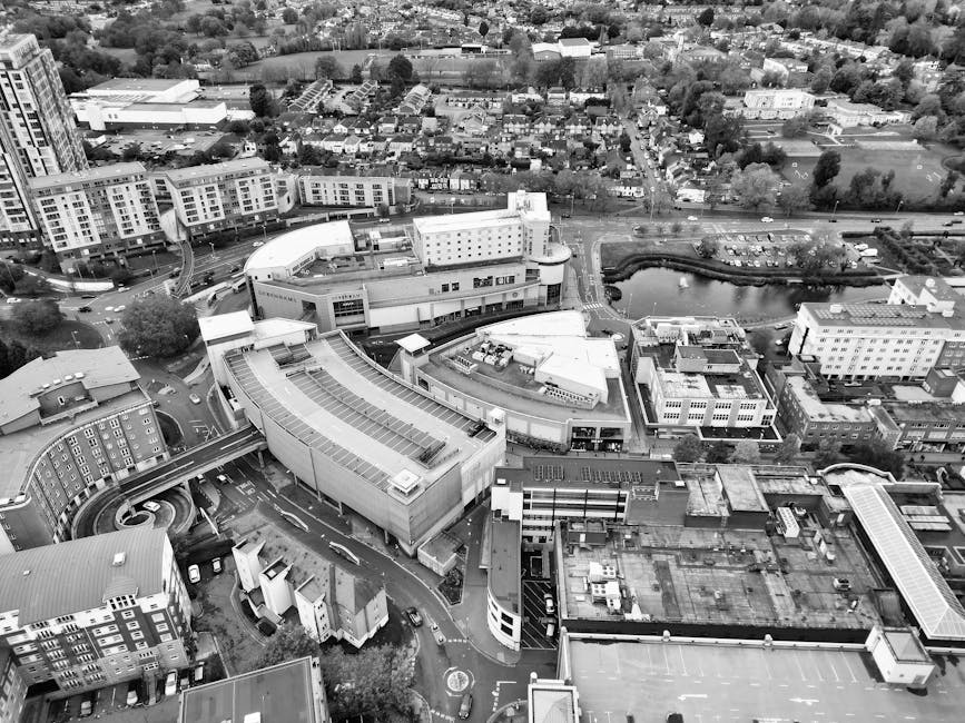 A black-and-white aerial photograph showing a busy urban area with multiple commercial and residential buildings, including a large modern multi-storey structure with a curved facade and a rooftop with skylights, likely part of a shopping centre or office complex. Surrounding the main building are smaller office buildings, some with flat roofs and rooftop equipment. The scene includes a network of roads with visible vehicles, some parked and others in motion, as well as a curved pedestrian walkway or bridge. In the background, there are rows of houses, green spaces, and a small pond surrounded by trees, indicating a mix of urban and suburban environments. This image reflects a dense cityscape typical of a town centre, with potential relevance to house removals, furniture transport, and moving logistics, supported by the context of [COMPANY_NAME] working in the area for relocation services.