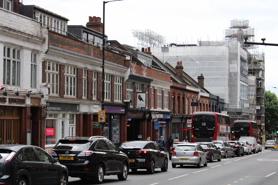 A street scene showing a row of brick and white-painted shopfronts and residential buildings along Station Road in Harrow, with some buildings undergoing construction and scaffolding on the right side. Several cars, including black, silver, and white vehicles, are parked along the curb, while others are driving or waiting at the roadside. A double-decker bus with route number 343 is visible, along with a nearby bus stop. The street is busy with pedestrians, some standing and others walking on the sidewalk. The scene is captured in daylight under overcast skies, highlighting typical city traffic and urban architecture, all relevant to a home relocation or furniture transport context as serviced by Man and a Van Harrow.