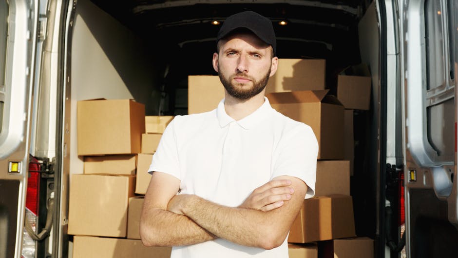 A young man with a serious expression, sporting a black cap and a white polo shirt, stands inside the rear of a moving van during a home relocation process. Behind him, the van's interior is visible, filled with neatly stacked cardboard moving boxes of various sizes, some sealed with packing tape. The boxes are arranged for optimal space utilization, indicating an organized loading process. The man is positioned near the open van doors on a paved surface, with natural light illuminating the scene. The image captures the loading area during daytime, reflecting the packing and furniture transport stages of a house removal, with visible equipment such as straps and blankets used to secure items. This scene illustrates a typical furniture transport operation coordinated by [COMPANY_NAME], highlighting the careful handling of belongings during same-day move preparations for services listed under removals.
