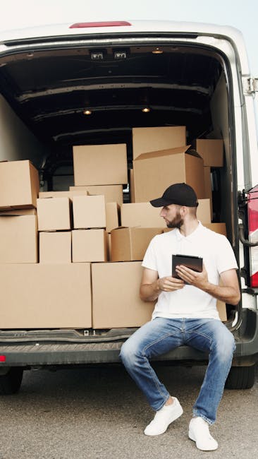 A man wearing a black cap, white t-shirt, and blue jeans is sitting on the pavement next to the open rear of a large white van, which is loaded with multiple cardboard boxes of various sizes, some sealed with packing tape. The boxes are stacked inside the vehicle, filling most of the space, and are prepared for a house move or furniture transport as part of a home relocation process. The man is holding a tablet device and appears to be checking or managing the packing and loading process. The van is parked on a street, with the surrounding environment not visible in detail. This scene illustrates the loading phase of a professional removals service, such as those offered by Man and a Van Harrow, focusing on efficient packing, transportation, and logistics for house removals.