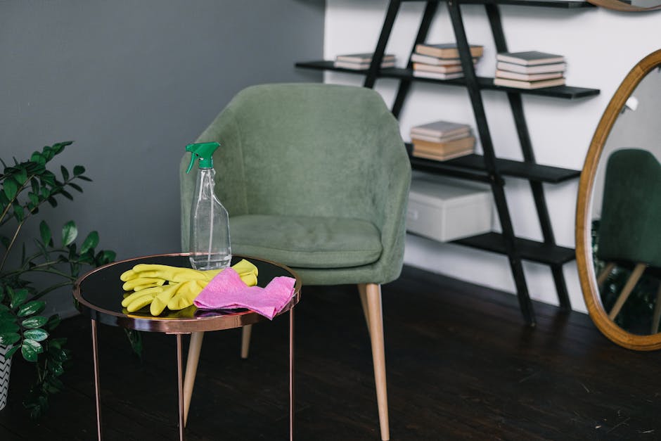 Inside a residential property, a cleaning spray bottle, yellow rubber gloves, and a pink cleaning cloth are placed on a small, round, dark-colored side table with metal legs. In the background, there is a light green, upholstered armchair with wooden legs positioned against a wall painted in a muted blue-grey shade. To the right, a black, modern-style bookshelf with multiple shelves holds a collection of books, some stacked horizontally, and a white storage box. A large oval mirror with a golden frame leans against the wall, reflecting part of the room, including another view of the armchair. The dark wooden floor provides a contrast to the furniture and cleaning supplies, suggesting a homemaking or post-removal cleaning process. This scene is relevant to house removals and furniture transport, often involving cleaning and preparation before or after moving items, which is occasionally managed by services like Man and a Van Harrow.