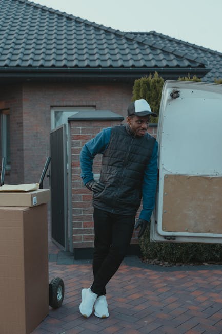 A man dressed in a blue long-sleeve shirt, black padded vest, and black trousers stands outside a residential property during a house removal process, leaning against a white moving van parked on a paved driveway. The van’s rear door is open, revealing some packing materials and a cardboard box on a trolley nearby, indicating furniture transport or packing activities. The man is wearing white protective footwear, commonly used during moving to prevent dirt tracking inside, and has a neutral expression while looking away. Behind him, there is a brick house with a dark tiled roof, a partially visible garden hedge, and a small pathway leading to the entrance. The scene is set in natural daylight, capturing the typical environment involved in home relocation with the focus on the loading process and packing materials associated with house removals, and illustrating the professional services provided by Man and a Van Harrow.