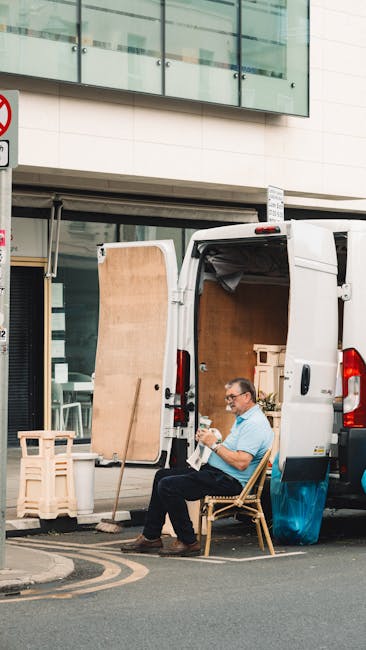 A man seated on a wooden chair beside a white delivery van parked on a city street, with the van's rear doors open revealing a large wooden panel, cardboard boxes, and other packing materials inside. He is holding a small object in his hands, possibly taking a break during a home relocation or furniture transport process. The scene is in front of a modern building with glass windows and a covered sidewalk area, with some plastic crates and a chalkboard sign nearby. The surrounding environment suggests an active loading area where packing and moving activities are underway, consistent with house removal services provided by Man and a Van Harrow.