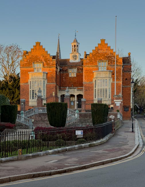 An exterior view of a historic red-brick building with a symmetrical facade featuring large bay windows, decorative brickwork, and a central clock tower with a weather vane on top. The building is situated behind a low stone wall and black wrought iron fencing, with a small landscaped garden area in front that includes neatly trimmed shrubs and flowering plants. A curved sidewalk runs along the edge of the property, adjacent to a quiet street with a lamppost visible on the right. The scene is illuminated by natural daylight, highlighting the architectural details and the surrounding trees, suggesting a peaceful neighbourhood setting suitable for residential or professional services such as house removals and moving logistics, as represented by local moving service providers like Man and a Van Harrow.