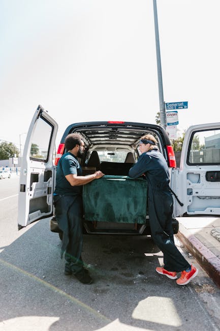 Two movers from Man and a Van Harrow, dressed in dark uniforms, are lifting and loading a large, green upholstered piece of furniture into the open rear cargo area of a white panel van parked on a city street. The van's interior is visible, showing shelving and a spacious compartment for packing household items. The scene takes place outdoors in daytime, with a clear sky and nearby street signs and pavement visible. The movers appear to be carefully managing the furniture, supporting its weight as part of a home relocation or furniture transport process, with the vehicle positioned close to the curb for efficient loading. The environment suggests a typical urban setting, reflecting the logistics involved in professional removals and packing services managed by Man and a Van Harrow.