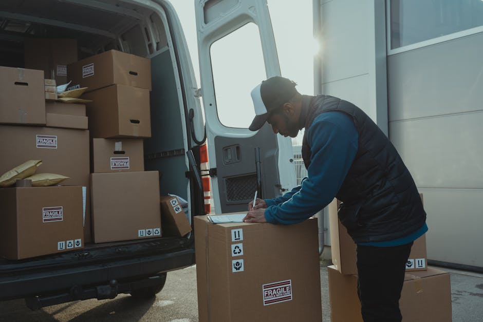 A man wearing a baseball cap, dark vest, and blue long-sleeve shirt is standing outside near the open rear of a white van, which is loaded with multiple cardboard boxes sealed with packaging tape. Some boxes have labels indicating fragility, and a few are wrapped in protective brown paper or plastic wrap. The man is writing on a clipboard placed on a box, likely recording inventory or moving details, as part of a home relocation or furniture transport process. The van's interior shows additional boxes stacked on the floor, ready for loading or unloading. The environment is well-lit with natural sunlight, and the van is parked on a paved surface adjacent to a building with grey paneling. This scene illustrates a professional moving or removals service, such as those provided by Man and a Van Harrow, during packing and loading activities for local house removals.