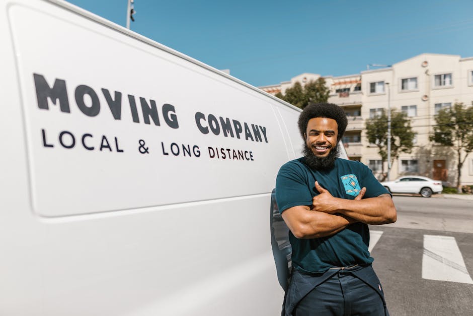 A smiling man with a beard and natural curly hair stands beside a white moving van on a city street, with residential buildings and parked cars visible in the background. The van features black text indicating it is a moving company offering local and long-distance services. He is wearing a dark blue t-shirt with a logo on the chest, and has his arms crossed in front of him. The scene is set outdoors during daytime with clear skies. The image captures the loading or unloading process associated with house removals, including the vehicle used for furniture transport and the professional appearance of the mover, as part of a home relocation or packing and moving service provided by Man and a Van Harrow.