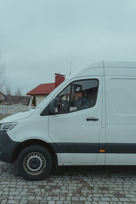 A white panel van parked on a paved driveway in a residential area, with a man seated inside the driver’s seat, visible through the open window. The man appears to be wearing a dark jacket and glasses. Behind the van, there is a house with a red roof, a white picket fence, and overcast sky with grey clouds. The scene suggests a house removal or furniture transport process, typical of professional relocation services like those offered by Man and a Van Harrow, involving loading or unloading household items during a flat removals in Harrow.