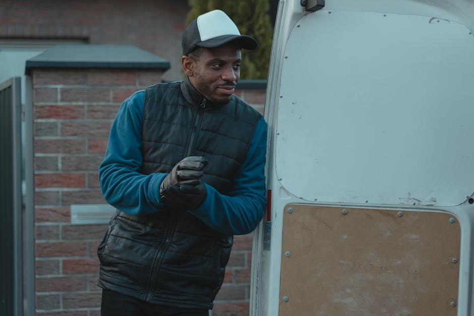 A man wearing a black and grey cap, blue long-sleeve top, and black gloves stands next to the open back of a white moving van, engaged in a home relocation process. Inside the van, visible are cardboard boxes, some wrapped in plastic, and other packing materials arranged near the loading area. The man is holding an object in his gloved hands, possibly preparing to load or unload items during furniture transport. The backdrop features a brick wall and greenery, indicating an exterior setting. The scene depicts a professional moving activity associated with house removals and packing and moving services, with the van positioned on pavement ready for transportation. Man and a Van Harrow, a company specialising in removals, appears to be coordinating the loading process in an organized manner, supporting efficient home relocation logistics.