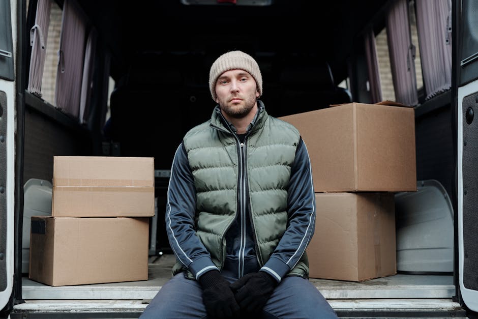 A man dressed in a grey beanie, a green and navy padded jacket, and black gloves is sitting inside the open cargo area of a van, which is lined with a grey interior and has its rear doors fully opened. Behind him are several cardboard boxes of various sizes, some stacked on top of each other, ready for moving or transport. The boxes appear sealed with packing tape and are likely used for packing household items during a home relocation. The van is parked on a paved surface, with the background indicating an outdoor setting. Inside the vehicle, the loaded items suggest an ongoing furniture transport or packing process, typical of professional removals services. The presence of packing materials and the arrangement of boxes highlight the loading process in preparation for a house move. Man and a Van Harrow’s team often handles similar setups during flat removals and residential relocations, as observed here.