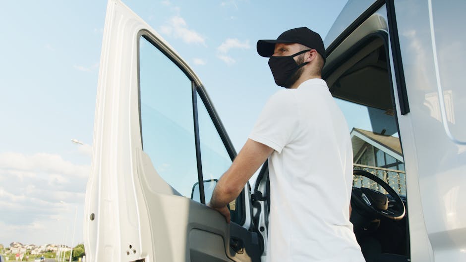 A man wearing a black face mask, black cap, and white T-shirt is standing inside the cab of a white moving van, holding onto the door frame as he prepares for a home relocation or furniture transport. The van is parked outdoors on a paved area with other residential buildings visible in the background under a partly cloudy sky. The interior of the van includes a steering wheel and dashboard, indicating the driver’s position. The scene suggests the loading process involved in packing and moving services provided by Man and a Van Harrow, with the focus on safety and logistics during house removals, likely including the handling of boxes, furniture, and packing materials inside the vehicle.