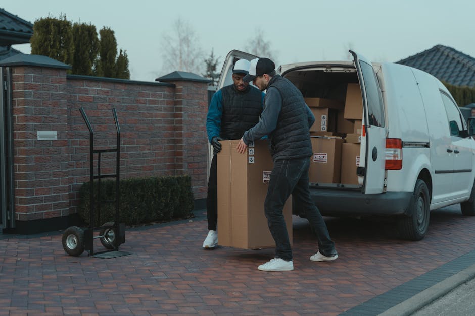 Two men are engaged in a home relocation process outside a residential property, loading a large cardboard box into the rear of a white delivery van, which is parked on a brick driveway near a brick wall and a hedge. One man, wearing a black beanie, dark vest, and grey pants, is lifting the box from the ground, while the other man, dressed in a black cap, black vest with blue sleeves, and black pants, is guiding it into the van. Inside the van, additional cardboard boxes can be seen organized on the floor, ready for transport. A black two-wheeled hand trolley stands nearby on the driveway, ready to assist with moving heavy items. The scene is set in overcast weather, with some trees and rooftops visible in the background, highlighting the logistics involved in furniture transport and packing for a flat move, with Man and a Van Harrow providing professional removals services.