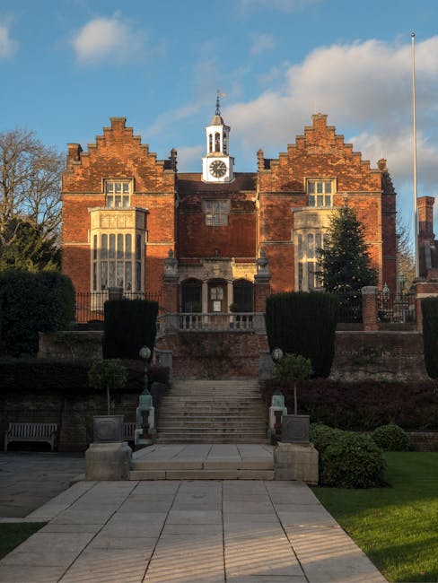 A historic red-brick building with a central clock tower featuring a white cupola and a spire, surrounded by a well-maintained garden and a stone staircase leading up to the entrance. The building’s façade includes large Gothic-style windows and decorative brickwork, with some areas darker due to weathering. In the foreground, there are paved walkways, trimmed bushes, and a park bench on the left side, indicating a landscaped outdoor area. The scene is illuminated by daylight, with a partly cloudy sky overhead. This classical structure, possibly a civic or educational building, is situated in a residential or institutional setting. For house removals and home relocation services, such as those provided by Man and a Van Harrow, understanding the exterior environment and access points is vital for planning furniture transport and loading processes.