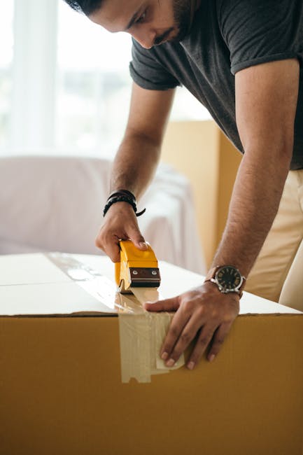 A man wearing a dark grey T-shirt and a wristwatch is using a yellow box cutter to remove packing tape from a large cardboard box. The scene appears to be inside a residential property, with natural light coming through a window in the background. The man is focused on preparing the box for opening or packing, with his hands firmly gripping the box and the cutter. The cardboard box is positioned on a surface, possibly a table or the floor, and is part of a house removal or moving process facilitated by Man and a Van Harrow, as suggested by the context of house removals and furniture transport. The environment is clean and well-lit, emphasizing the careful handling of packed items during a home relocation.