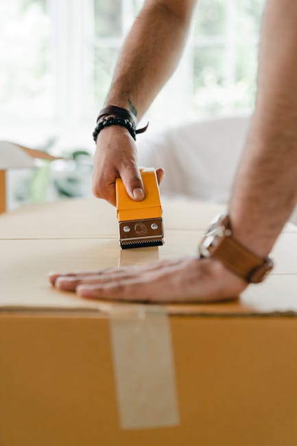 Close-up of a person's hands during a home relocation, with one hand pressing down on a large cardboard box wrapped in clear packing tape, while the other hand holds a yellow and silver box cutter or utility knife, cutting or sealing the tape. The individual wears a black wristband and a watch, with a bright, well-lit interior space in the background, featuring large windows and greenery outside. This scene illustrates packing and moving activities typical of furniture transport and home removals, with the focus on careful preparation of packages for loading onto a moving vehicle, supported by the professional services of Man and a Van Harrow.