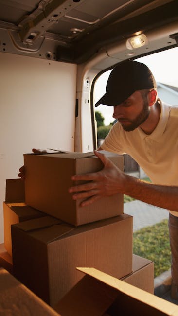 A man wearing a black cap and a beige polo shirt is inside the back of a van, carefully stacking cardboard moving boxes. The boxes are of various sizes, some wrapped in plastic or packing materials, and are arranged on the vehicle's floor near the open rear door. The interior of the van features metal walls and ceiling components suitable for furniture transport and packing during a home relocation. Outside the van, a grassy area and some trees are visible in the background, indicating daylight and outdoors. This scene depicts the loading process typical of house removals and furniture transport, with the man managing packed belongings ready for a removal service provided by Man and a Van Harrow, supporting the logistics of student removals in Harrow.
