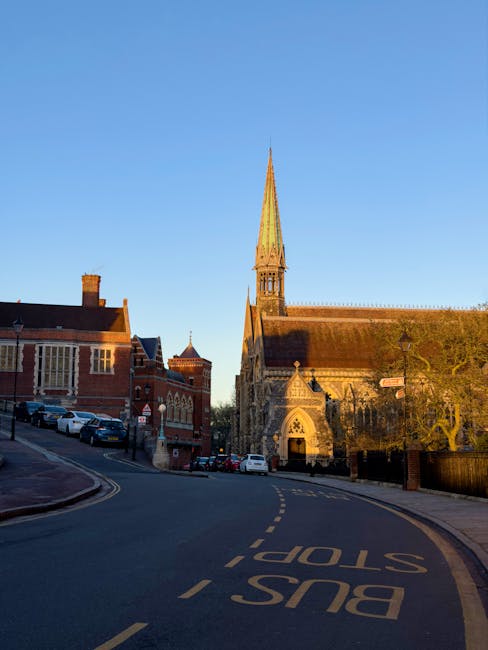 A street scene featuring a historic church with a tall, pointed spire and detailed stonework, situated alongside red-brick buildings with traditional architectural elements. The street has a gentle curve with a yellow dashed line in the middle and a prominent 'BUS STOP' marking painted on the tarmac, indicating the area designated for public transportation. Several cars are parked along the roadside, including a mix of small and medium-sized vehicles, and a few are in motion, suggesting an active urban environment. The scene is captured during daytime under clear blue skies, with sunlight illuminating the facades of the buildings and casting soft shadows. This setting could relate to a suburban or town centre location where home relocations and moving activities, such as those handled by Man and a Van Harrow, may take place near notable landmarks or in residential streets close to local services.