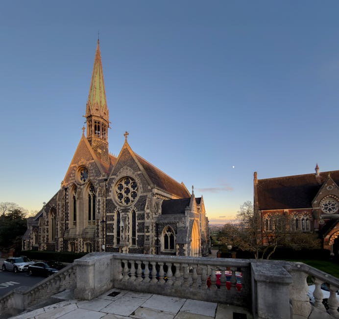 A large, historic stone church with intricate Gothic architecture, featuring tall pointed spires, large arched stained glass windows, and detailed stone carvings. The church is situated outdoors, with a clear sky in the background, and the setting appears to be either early morning or late afternoon, as indicated by the soft, warm sunlight. In the foreground, there is a stone balustrade or terrace with a weathered surface, overlooking the church. To the right of the church, a smaller building with similar architectural features is visible, with additional stained glass windows and a pitched roof. The scene captures a peaceful, well-maintained area, suitable for home relocation or moving services, with no visible vehicles or moving equipment present in this image.