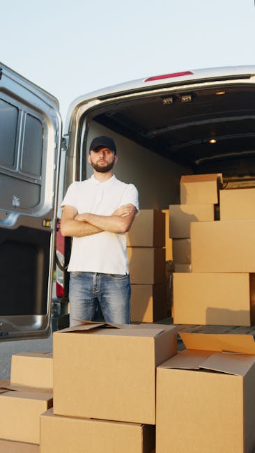 A man with a beard, wearing a dark cap, white T-shirt, and jeans, stands with arms crossed beside an open van during a home relocation process in daylight. The van's interior is visible and filled with stacked cardboard boxes of various sizes, some with flaps open, prepared for packing and moving. Several boxes are positioned on the pavement in front of the van, ready for loading or unloading. The background shows a clear sky, indicating outdoor activity in a residential or urban area. This scene illustrates furniture transport and packing procedures typical of professional removals services, with Man and a Van Harrow waarschijnlijk betrokken in coordinating the loading process for a house or flat removal, as referenced in the page titled 'Flat Removals Harrow' on [DOMAIN].