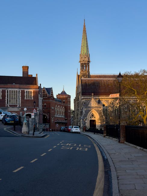 A view of a street outside a historic church with a tall, pointed spire, situated on a corner with a brick building to the left and trees to the right. Several parked cars are visible along the pavement, and a traffic island marks the bus stop area on the road. The street has dashed white lines and a sign indicating a bus stop, with a lamppost nearby. The scene is captured during daylight with clear blue skies, and the overall setting suggests a quiet residential or town centre area. This urban environment relates to house and flat relocations, often involving moving logistics and furniture transport near buildings like this church. Man and a Van Harrow offers removals and moving services that could include handling furniture and household items in such locations.
