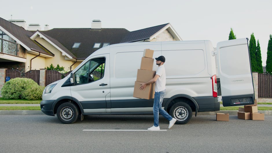 A man wearing a white t-shirt, blue jeans, and a black cap is standing on the street beside a white cargo van with its side door closed and the rear door open, in the process of loading or unloading furniture and boxes. He is holding three stacked cardboard boxes, with additional boxes on the ground nearby, some partially inside the van. The van is parked on a residential street in front of modern houses with pitched roofs, large windows, and balconies, surrounded by greenery including shrubs and tall trees. The scene depicts a home relocation or furniture transport operation, with the man and a van from Man and a Van Harrow engaged in packing or unpacking. The lighting suggests daytime with clear weather.
