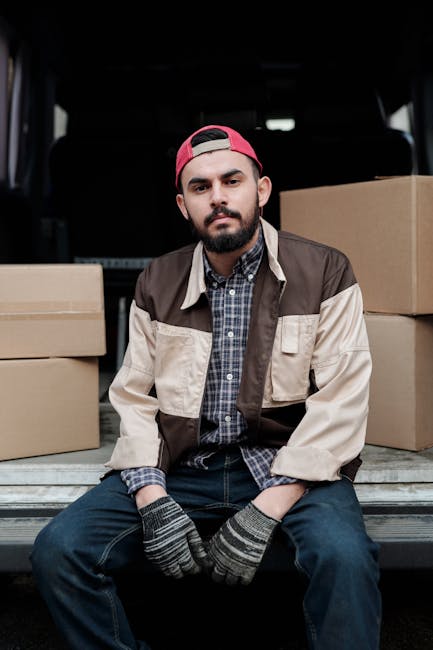 A young man with a serious expression, wearing a red cap, a checked shirt, a beige and brown jacket, and striped gloves, is sitting on the open tailgate of a black van during a home relocation process. Behind him, the van's interior is partially visible, with several cardboard boxes stacked on either side of him, some wrapped in plastic or packing material. The boxes vary in size and are positioned on a paved surface outside a property, with the van ready for loading or unloading. The scene depicts the packing and furniture transport aspect of house removals, with the man preparing to move belongings as part of a professional moving service provided by Man and a Van Harrow, supporting the logistics of student removals in Harrow.
