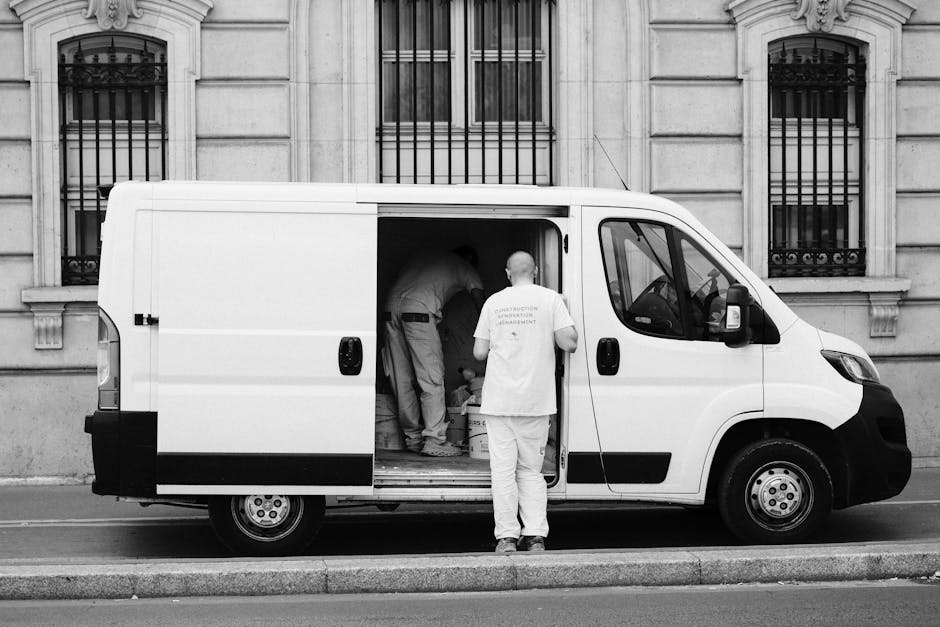 A white commercial van parked on a city street with its sliding side door open, revealing two movers inside handling packing materials and cardboard boxes as part of a furniture transport and home relocation process. One mover, dressed in light clothing, stands outside the van facing inwards, observing or waiting, while two other movers inside are actively lifting or arranging items on the van's floor, which is lined with protective blankets. The van is positioned adjacent to a stone building with decorative window bars, and the scene demonstrates the loading process involved in house removals or student moves, with equipment such as moving blankets visible to safeguard furniture during transit. The environment is illuminated by natural daylight, highlighting the urban setting where precise packing and careful loading are taking place, supported by professional removal services like Man and a Van Harrow.