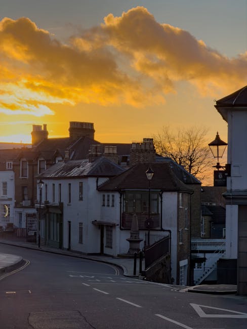 A street scene during sunset shows a row of residential buildings with a mix of brick and white facades, some with bay windows and chimneys, situated along a gently curved road marked with white lines and parking restrictions. In the foreground, a black metal railing lines a staircase leading down from a building on the right, which features a lamppost mounted on its corner. The background displays a vibrant orange and yellow sky with large, fluffy clouds illuminated by the setting sun. This peaceful urban setting may be part of a home relocation or moving process, typical of the services provided by Man and a Van Harrow, who specialise in removals and furniture transport across Harrow.