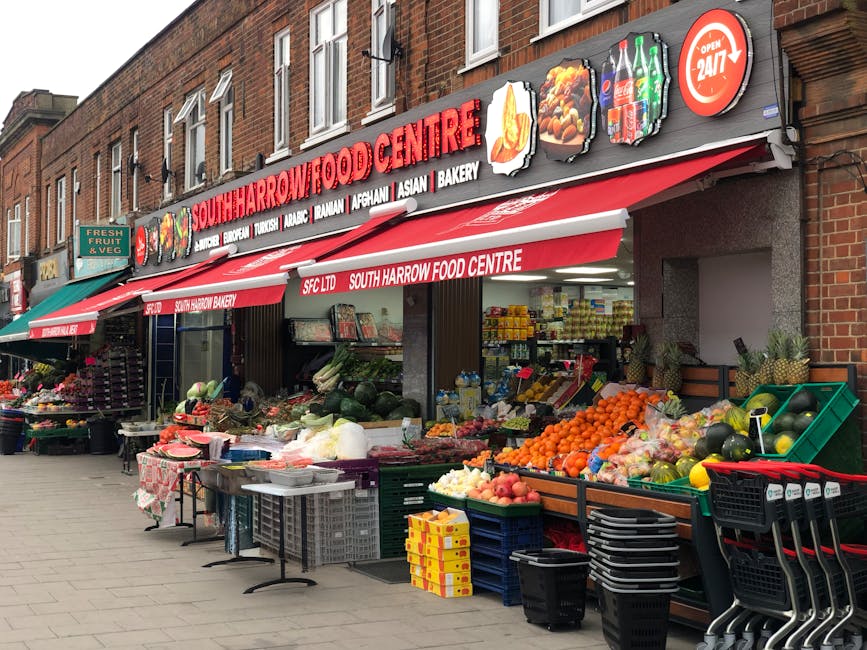 The image depicts the storefront of South Harrow Food Centre, a local grocery shop situated on a brick building with large windows and red signage displaying the shop's name. In front of the shop, there are outdoor displays of fresh produce arranged on green and black plastic crates, including oranges, watermelons, apples, cabbages, and other fruits and vegetables. Some produce is covered in plastic bags or wraps, and additional items such as bottled drinks, paper cartons, and packaged goods are visible inside the shop through the open entrance. To the left, there are small tables with more food items and a stall with flowers or plants. The pavement outside is made of paving stones, and the setting appears to be during daytime under natural lighting. This scene could be relevant to home relocation or packing activities, as South Harrow Food Centre may be a location where items are sourced for moving preparations, and Man and a Van Harrow offers removals and furniture transport services across the area.