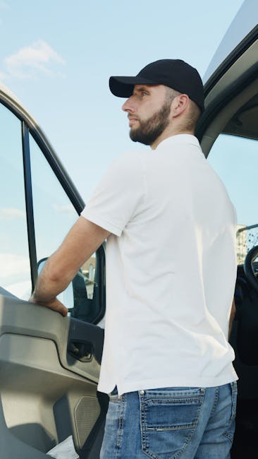A man with a beard, wearing a black cap and a white t-shirt, is standing next to the open door of a moving van, possibly preparing for a home relocation. Inside the vehicle, there are visible cardboard boxes, some wrapped in plastic or fabric, as well as furniture pieces such as a wooden table and other packed items. The background shows a clear sky with a few clouds, indicating daytime, and the van appears to be parked outside a property. The scene captures the loading process involved in furniture transport and packing during house removals, with the focus on the man readying belongings for transportation, illustrating aspects of a professional moving service offered by Man and a Van Harrow for flat removals in Harrow.
