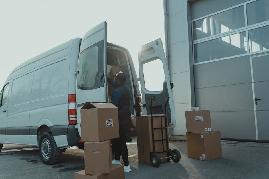A professional mover from Man and a Van Harrow is loading household boxes and packing materials into the back of a white cargo van during a home relocation. The mover, dressed in dark work clothing and a cap, is placing cardboard boxes with labels into the van, which has its rear doors wide open, revealing additional packed items inside. Several other cardboard boxes, some sealed with tape and others open, are positioned on the pavement nearby. A hand truck or dolly is standing upright next to the open van, ready to transport additional packing supplies or furniture. The setting appears to be the exterior of a modern building with large windows and a grey facade, during daylight with the sun visible in the background. This scene captures the loading process typical of furniture transport and packing during house removals, illustrating the logistical aspects of professional moving services provided by Man and a Van Harrow.