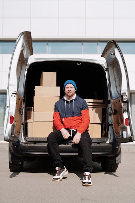 A young man with a blue beanie, navy and red jacket, and black pants is seated on the ground in front of the open rear doors of a white cargo van parked outside a modern building with a glass facade. The van's interior is filled with neatly stacked cardboard boxes, varying in size, some wrapped in plastic or protective packaging. The man appears to be smiling, with a relaxed posture, and the setting suggests he is involved in a home relocation or furniture transport activity. The loading area beneath the open van doors is clear, with a paved surface in the foreground. The scene captures the process of packing and moving, supported by Man and a Van Harrow's professional removals services, highlighting safe handling and transportation of packed household items during a residential move.