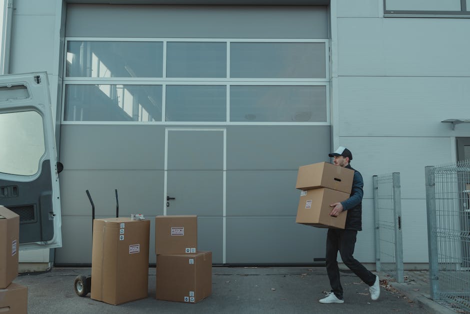 A man wearing a dark jacket, black trousers, white sneakers, and a baseball cap is moving multiple cardboard boxes, some stacked, during a house relocation or furniture transport task outside a modern industrial building with large glass windows and grey paneling. The boxes are of various sizes and are placed near a white panel van with its rear door open, revealing more packed boxes inside. On the ground, there is a metal hand truck and additional boxes awaiting loading. The man is carrying two boxes, one on top of the other, in preparation for transfer into the van or building. The scene captures the loading process involved in packing and moving services, with the man carefully handling the boxes in an organized manner. The setting is outdoors, with a paved surface and a metal fence adjacent to the building, under natural daylight conditions, illustrating a typical step in home relocation or furniture transport managed by Man and a Van Harrow.