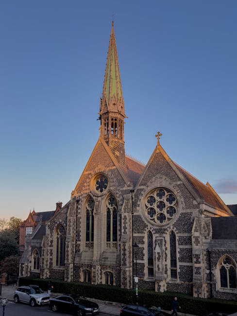 A historic stone church with Gothic architectural features, including pointed arches, intricate tracery windows, and decorative quatrefoil designs, situated on a street with parked cars and a hedge in front. The church has a tall, slender spire topped with a cross, and its exterior walls display a mix of flint and stone masonry. The scene is captured during the late afternoon or early evening with clear blue sky and soft sunlight illuminating the building. This image reflects the types of architectural structures involved in moving and relocation services, such as those provided by Man and a Van Harrow, which may include the careful transportation of such structures or related furnishings during house removals in the Harrow area.