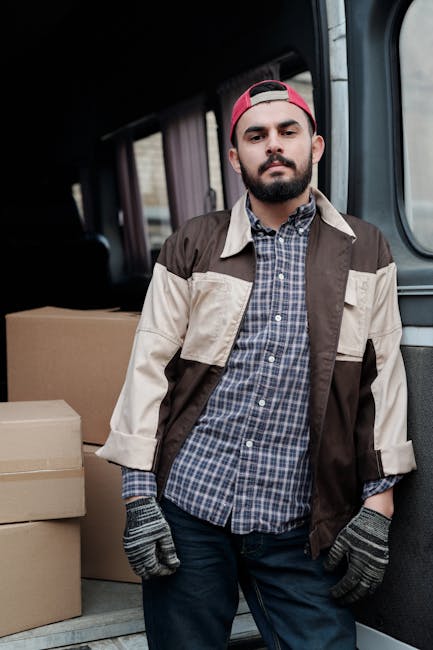A young man with a beard and wearing a red baseball cap, a beige and brown jacket over a blue checkered shirt, and grey work gloves, standing inside a van used for house removals. The van's interior shows a few cardboard boxes placed on the floor behind him, ready for packing or transportation during a home relocation. The background includes slightly visible interior elements like seats or shelving, with natural lighting illuminating the scene. This image depicts the logistics and staff involved in furniture transport and packing during a house removal service, aligning with professional moving processes by Man and a Van Harrow, specialists in house and student removals in Harrow.