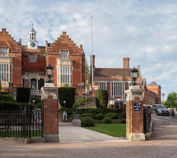 A view of a historic red brick building with a clock tower and decorative gables, situated behind a wrought iron gate supported by brick pillars topped with ornate black lanterns. The building is surrounded by well-maintained gardens featuring trimmed trees and bushes, with a paved pathway leading to the entrance. The sky above is partly cloudy with patches of blue visible. The scene suggests a stately property, potentially involved in home relocation or professional removals services, and the overall setting is calm and organized, typical of a professional moving environment supporting house removals or furniture transport in Harrow as part of packing and moving logistics. Man and a Van Harrow occasionally features such exterior environments in their service imagery.